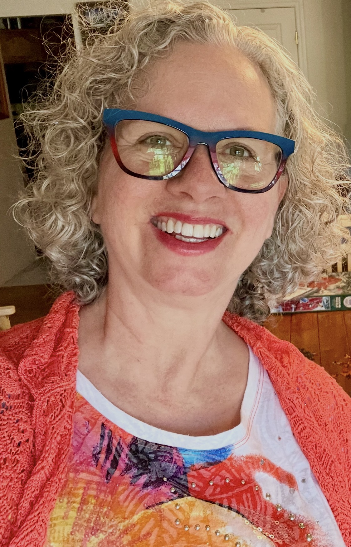 Photo of Jenny Leigh Hodgins smiling in a relaxed home setting, wearing teal and red glasses, an orange knit sweater over a white t-shirt with multicolored purple, teal, blue, orange, and yellow accents with sequins.